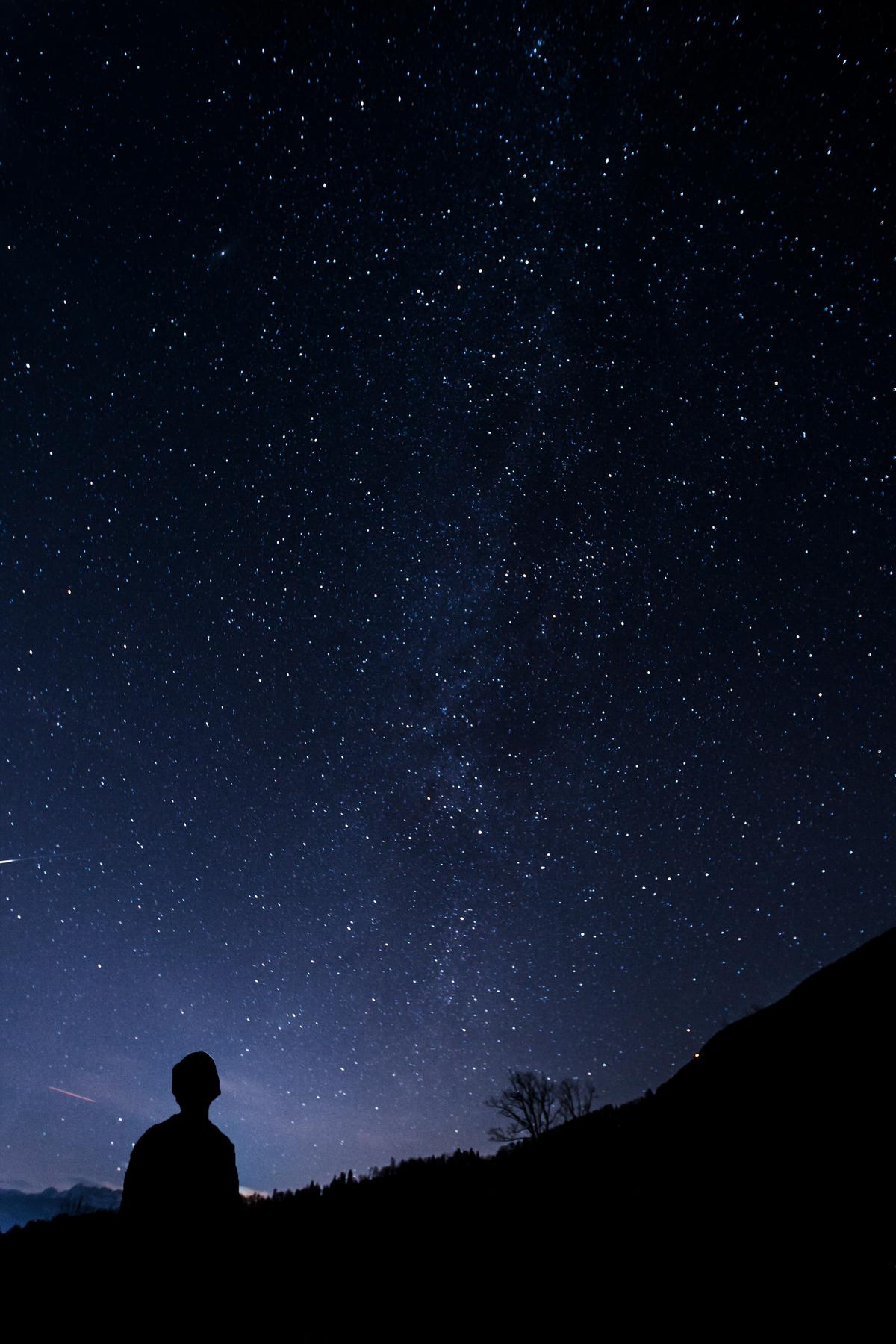 A hiker looking up at the night sky with a portable telescope, surrounded by mountains and stars.