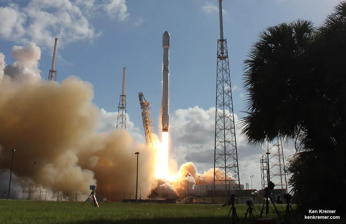 An aerial view of a SpaceX launch facility, showing multiple launch pads, support structures, and a rocket being prepared for launch